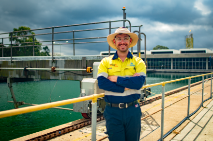 A man in a wide brimmed hat and hi vis clothing stands proudly in front of a long basin filled with water at a water treatment plant