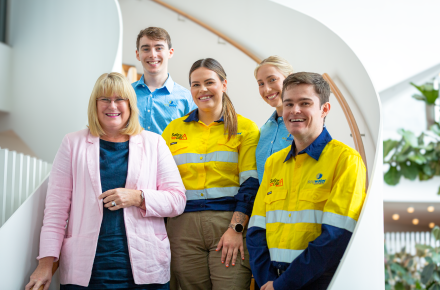 Minister Leahy with blonde hair and a pick jacket stands on a staircase with four young graduates in hi vis